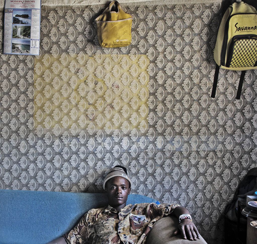 Un homme noir est installé sur un canapé bleu. Au mur un papier peint et des sacs jaunes accrochés.