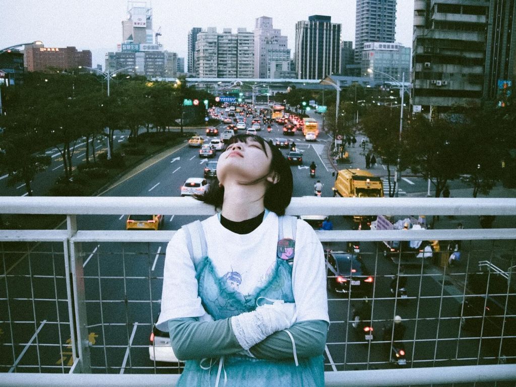 Une jeune femme, les bras croisés, pose sur un pont. En arrière-plan les voitures circulent