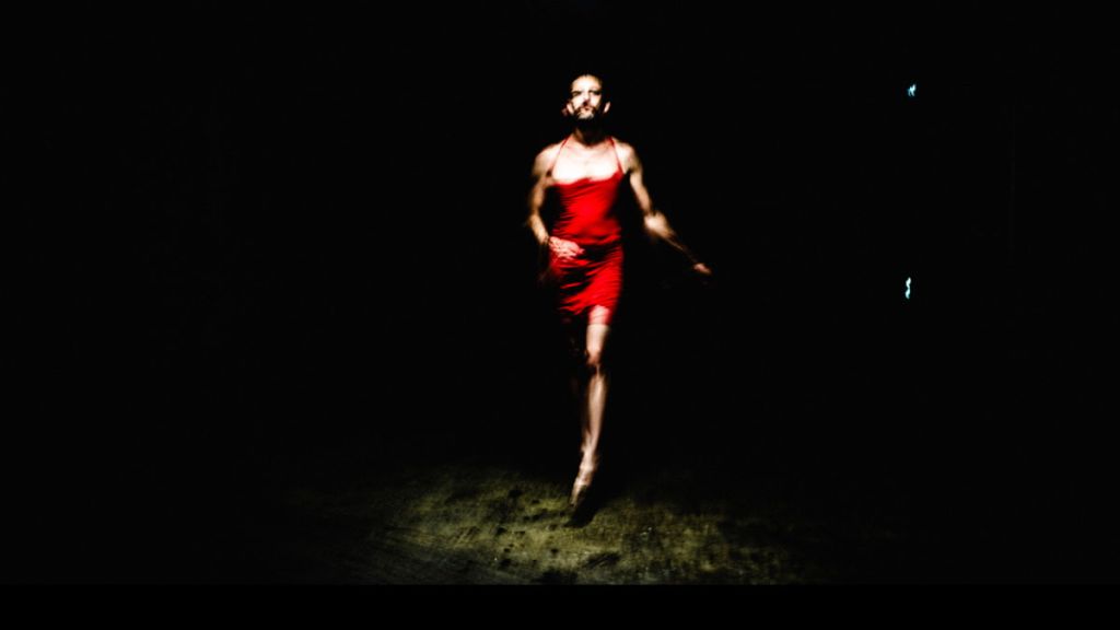 Une personne en petite robe rouge se tient debout face à l'objectif, dans une ambiance sombre