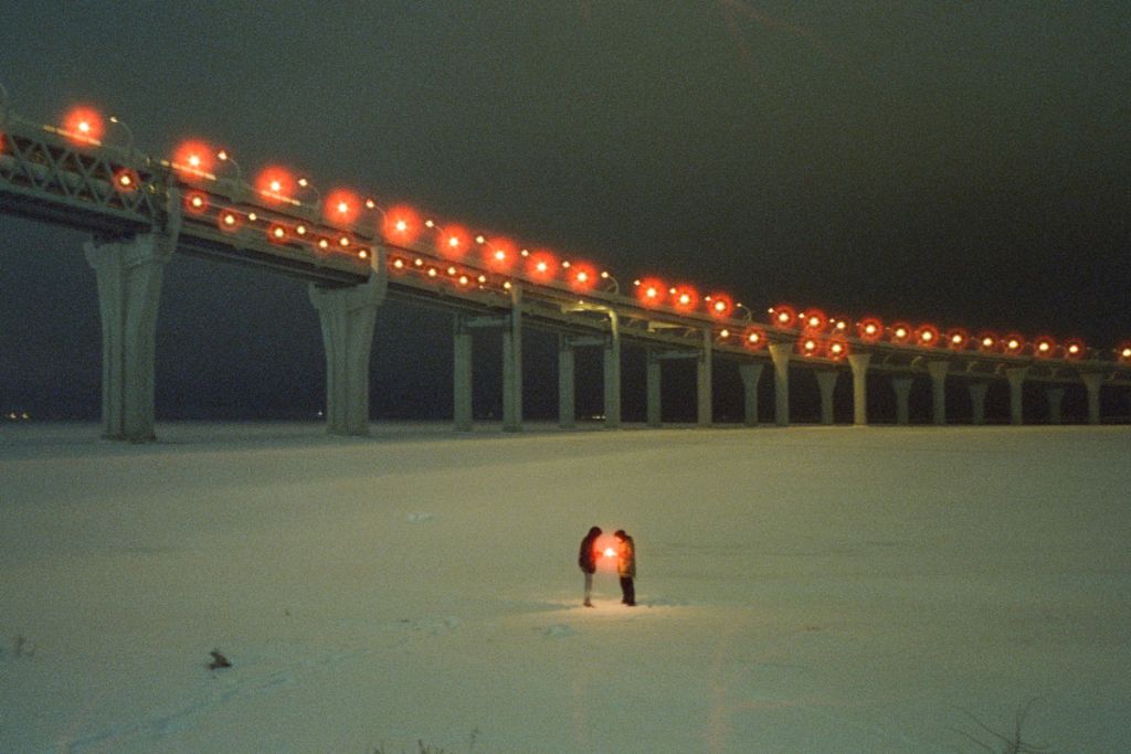 Deux silhouettes rapprochées se font face dans un paysage enneigé, sous un pont illuminé