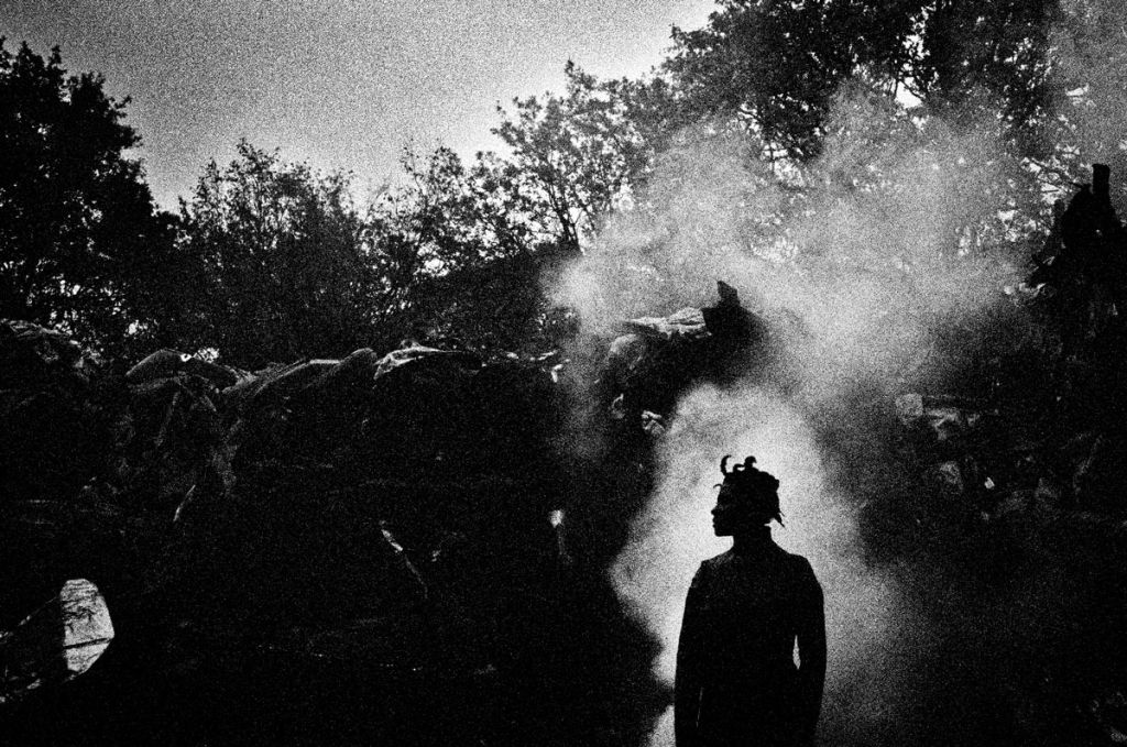 Photo en noir et blanc, la silhouette d'une figure de profil se tient au premier plan, derrière un tas d'objets semblables à une décharge d'ordures.