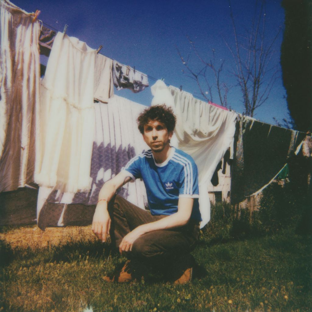 Le musicien Hannah Miette pose accroupi devant du linge blanc suspendu dans un jardin, portrait Polaroïd.