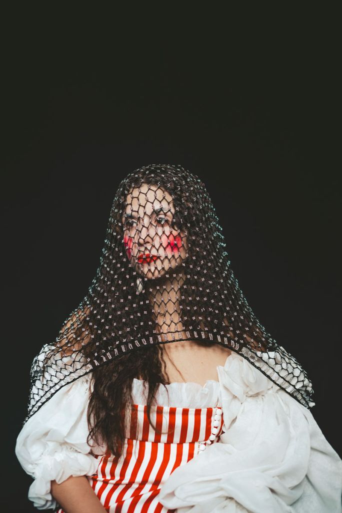 Portrait de la musicienne Helene Escriva, qui porte un maquillage de clown et pose dans un costume blanc aux rayures rouges.