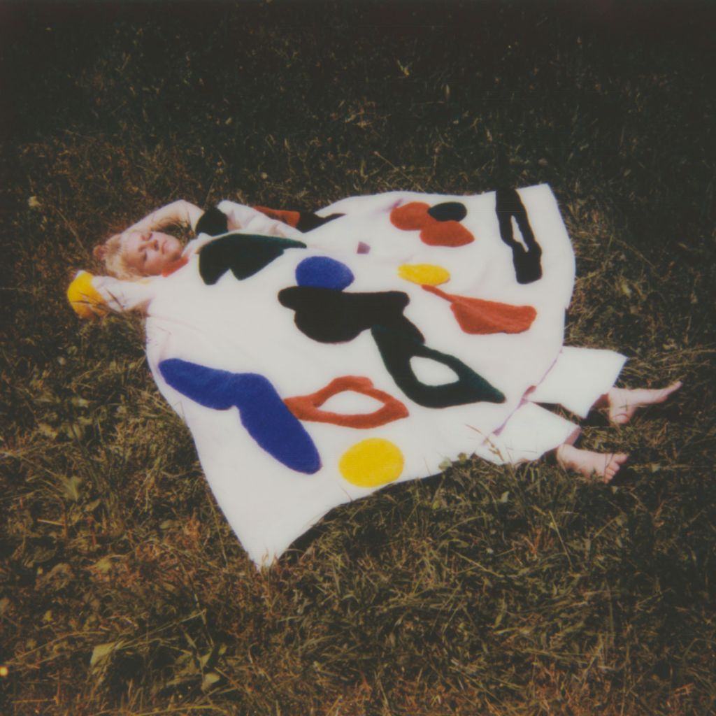 La musicienne française KCIDY est allongée au milieu de l'herbe, dans un costume coloré aux allures clownesques. Polaroïd.