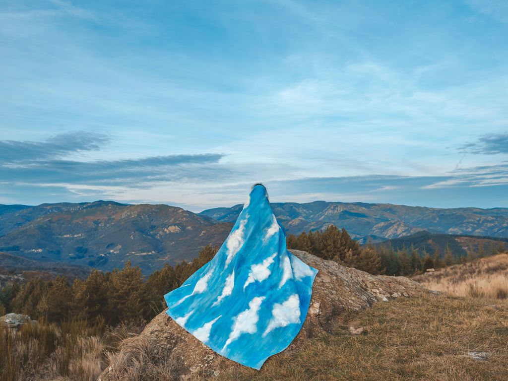 Une figure enveloppée d'un long tissu bleu et blanc trône de dos sur un rocher, face à un paysage de montagnes.