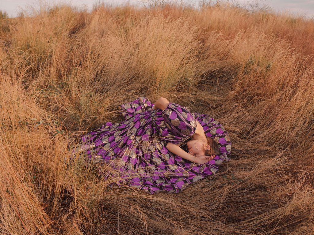 Une jeune femme en robe mauve allongée et recroquevillée sur elle-même, au milieu des hautes herbes.