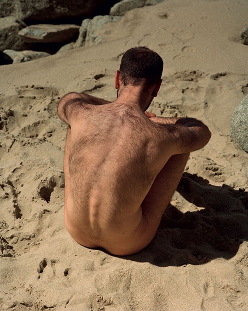 Photographie de Daniel Jack Lyons montrant un homme nu, de dos, assis sur le sable