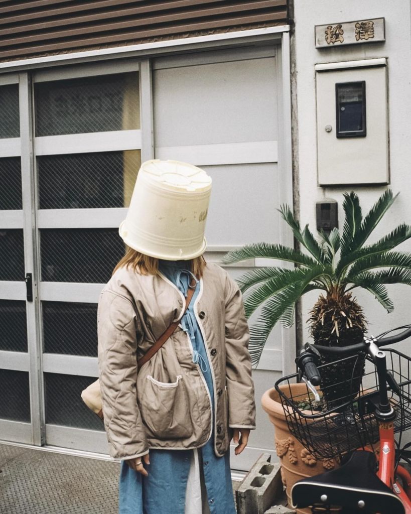 Une personne est debout dans la rue avec un seau sur la tête. À côté, une plante verte