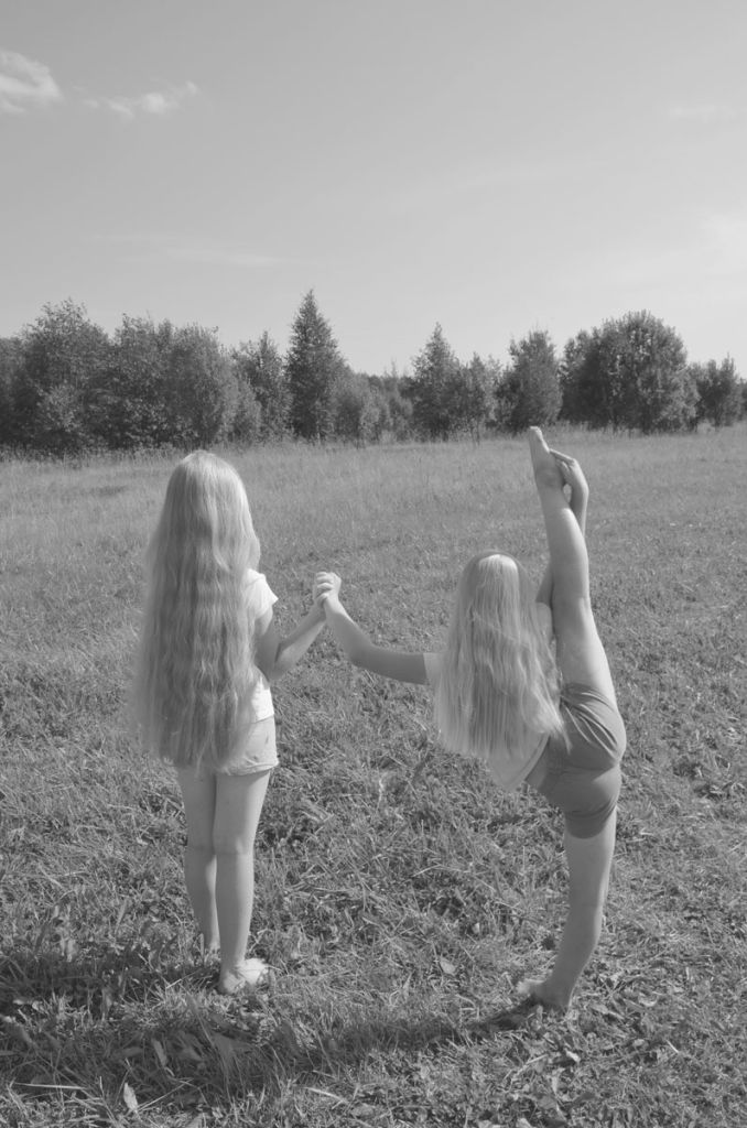 Photo en noir et blanc de deux jeunes filles blondes, de dos, aux cheveux longs, qui font face à une plaine. L'une d'elle étire l'une de ses jambes à la manière d'une ballerine, et s'appuie sur son amie pour tenir en équilibre.