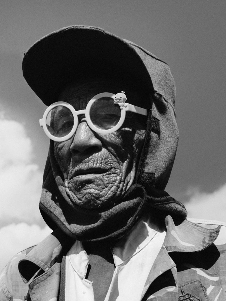 Image en noir et blanc d'une personne âgée, la tête recouverte d'un tissu et portant de grosses lunettes de vue blanches