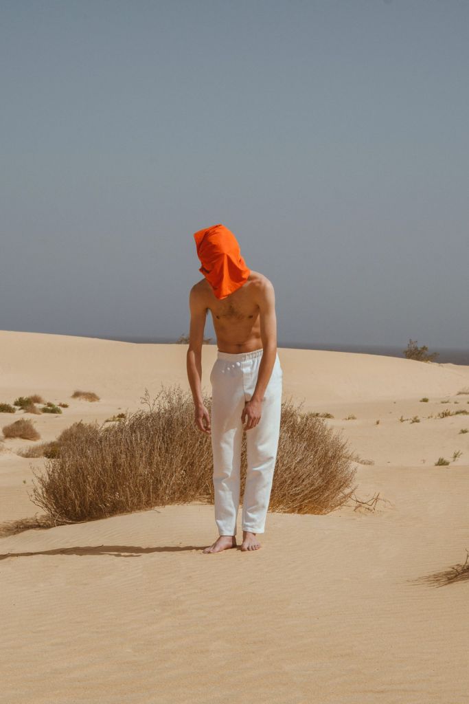 Personne vêtue d'un pantalon blanc, d'un foulard orange couvrant le visage sur une étendue de sable